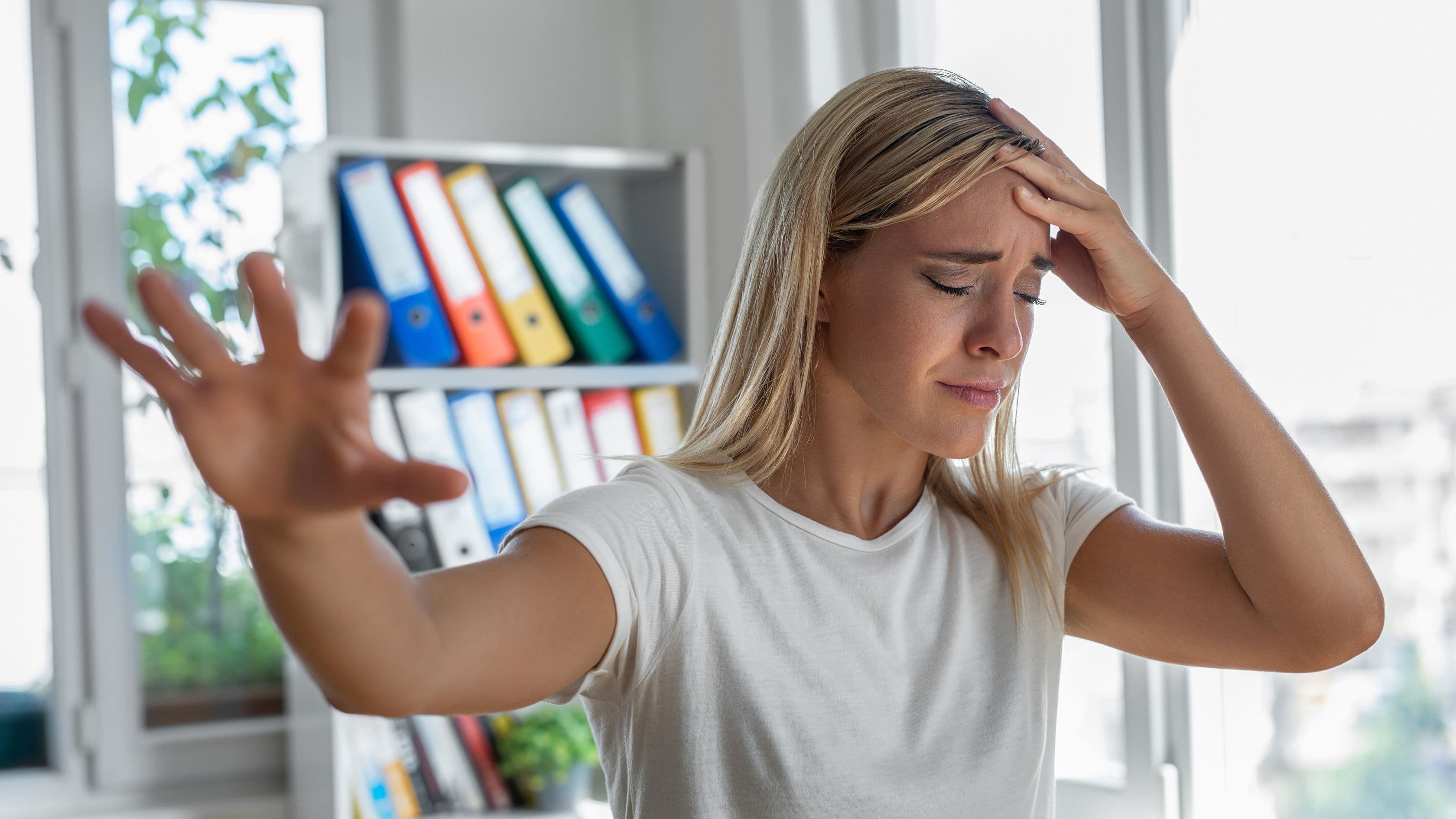 a woman is holding her head in pain while sitting at a desk in front of a window .