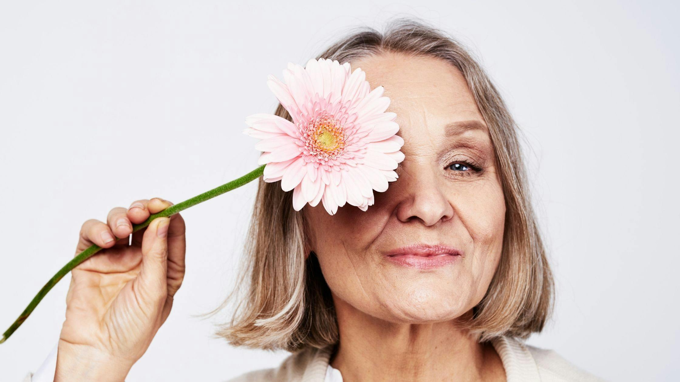 a woman is holding a pink flower in front of her eye .