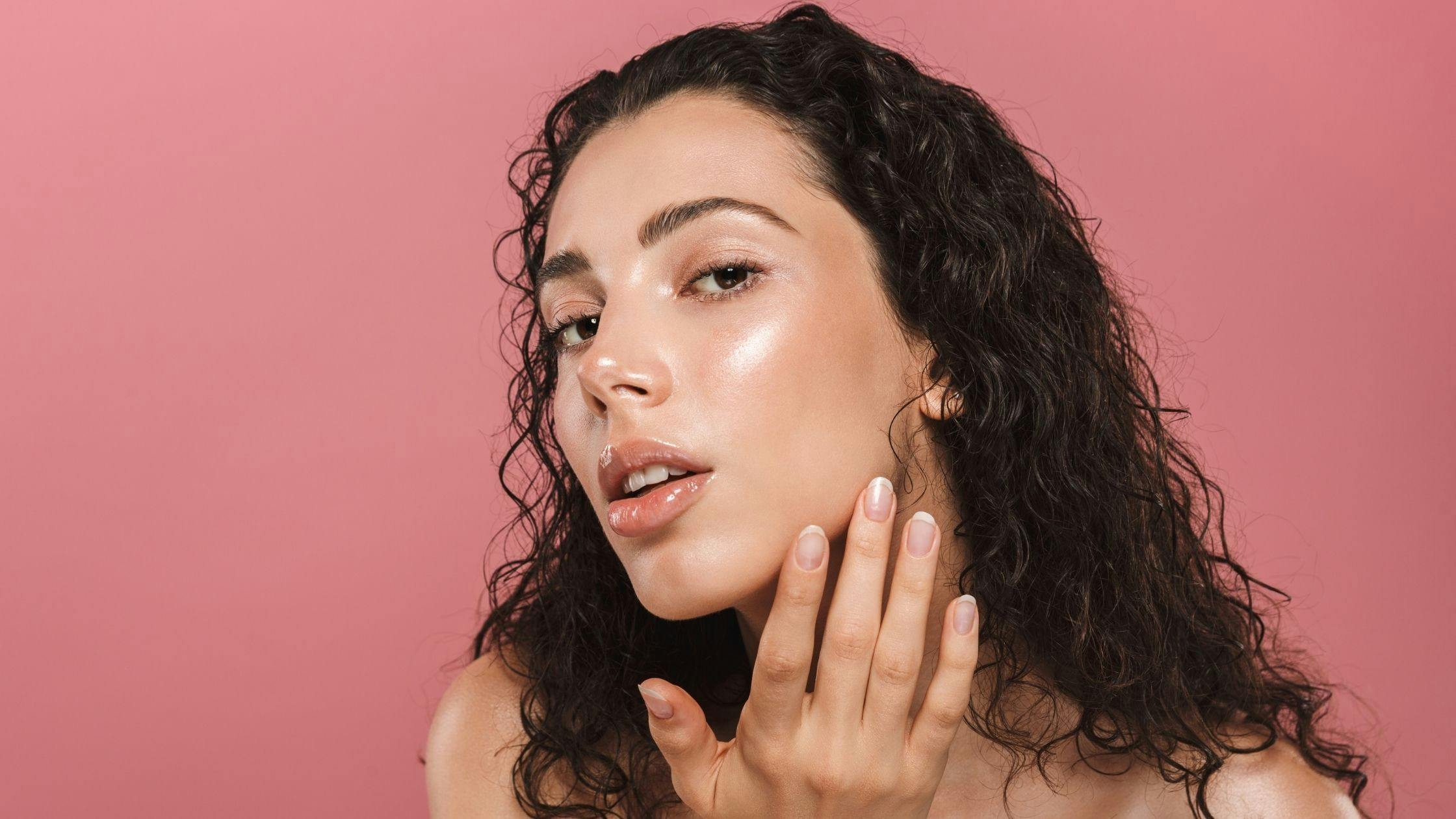 a woman with curly hair is touching her face on a pink background .
