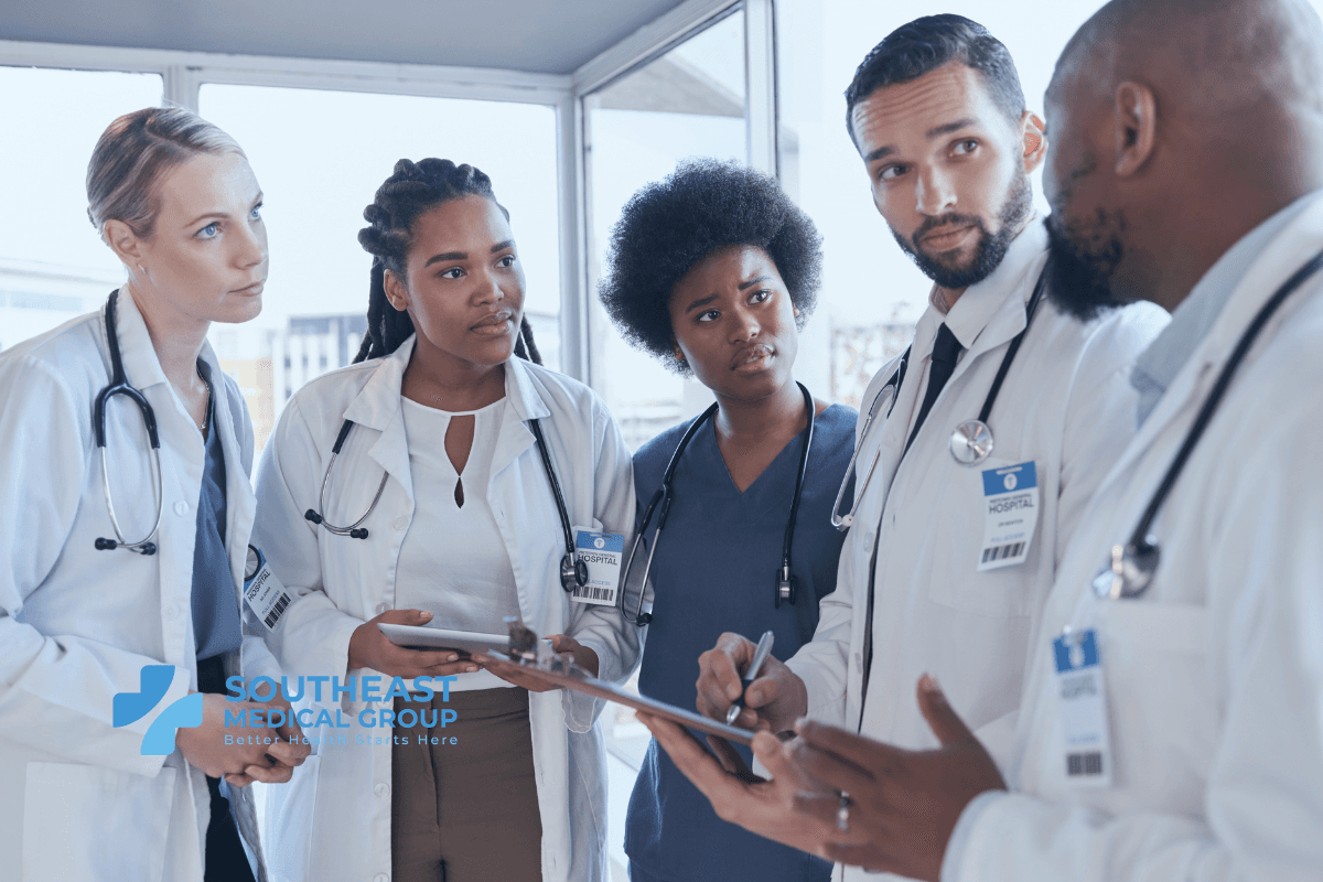 A diverse group of five medical professionals in lab coats and scrubs intently discussing and holding clipboards.