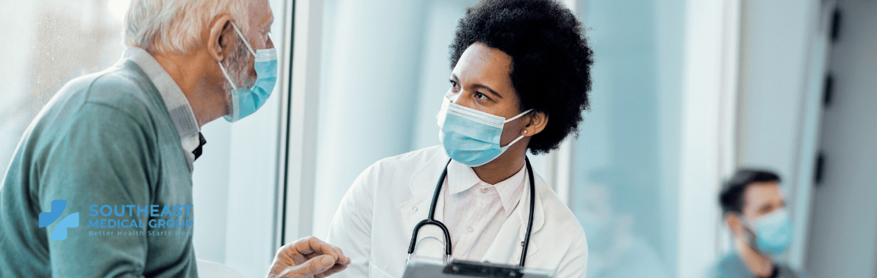 A Black female doctor in a mask talks to an older male patient in a mask.
