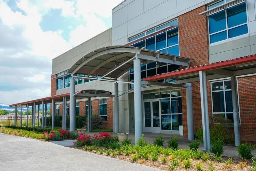 A modern brick and panel building with blue-tinted windows, a curved covered entrance, and a landscaped walkway.
