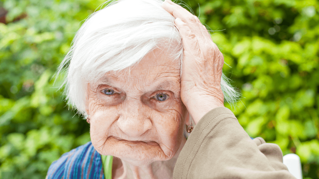 Elderly woman with white hair holding hand to her head, looking distressed.