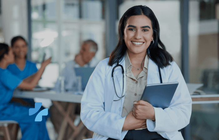 A smiling doctor with a stethoscope holds a tablet, with blurred medical professionals in the background and a blue logo.