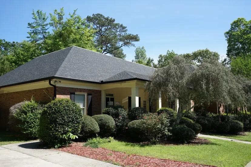 A single-story brick building with a dark roof, covered porch, and extensive green landscaping under a clear sky.