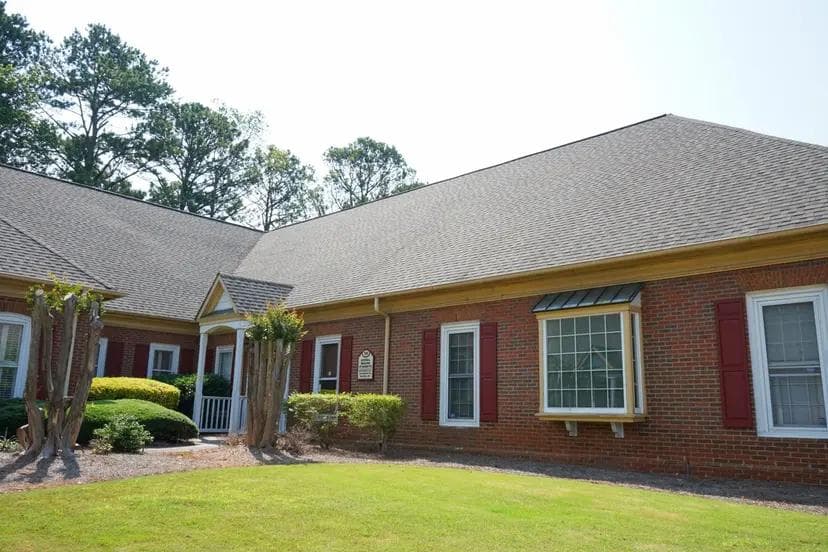 A brick building with a gray shingled roof, red shutters, and a bay window, with green grass in the foreground.