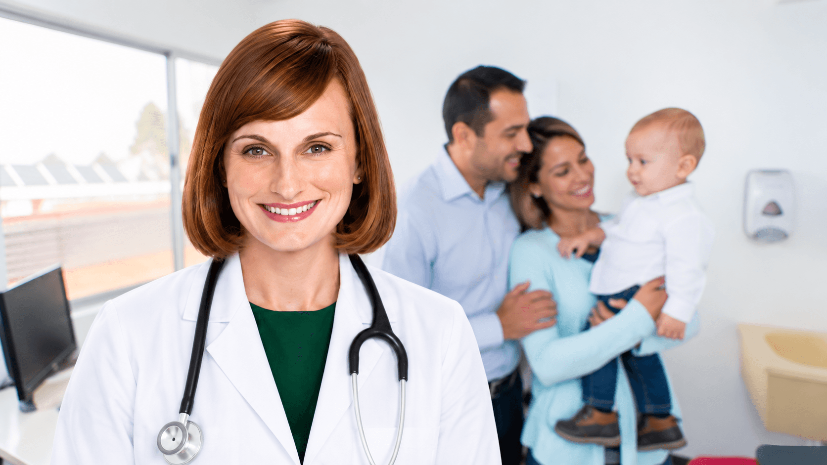A smiling female doctor with a stethoscope, with a family holding a baby in the background.
