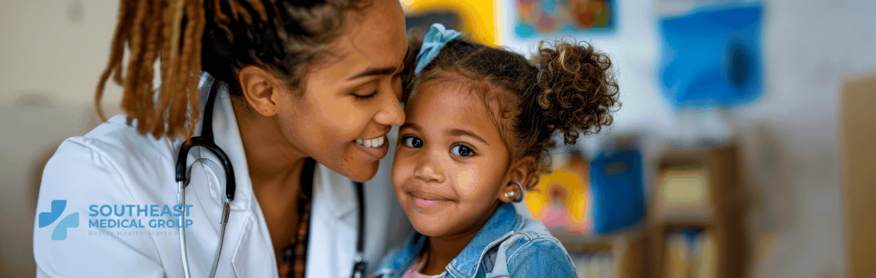 A smiling Black female doctor with a stethoscope leans toward a smiling young Black girl. The Southeast Medical Group logo is on the doctor's coat.
