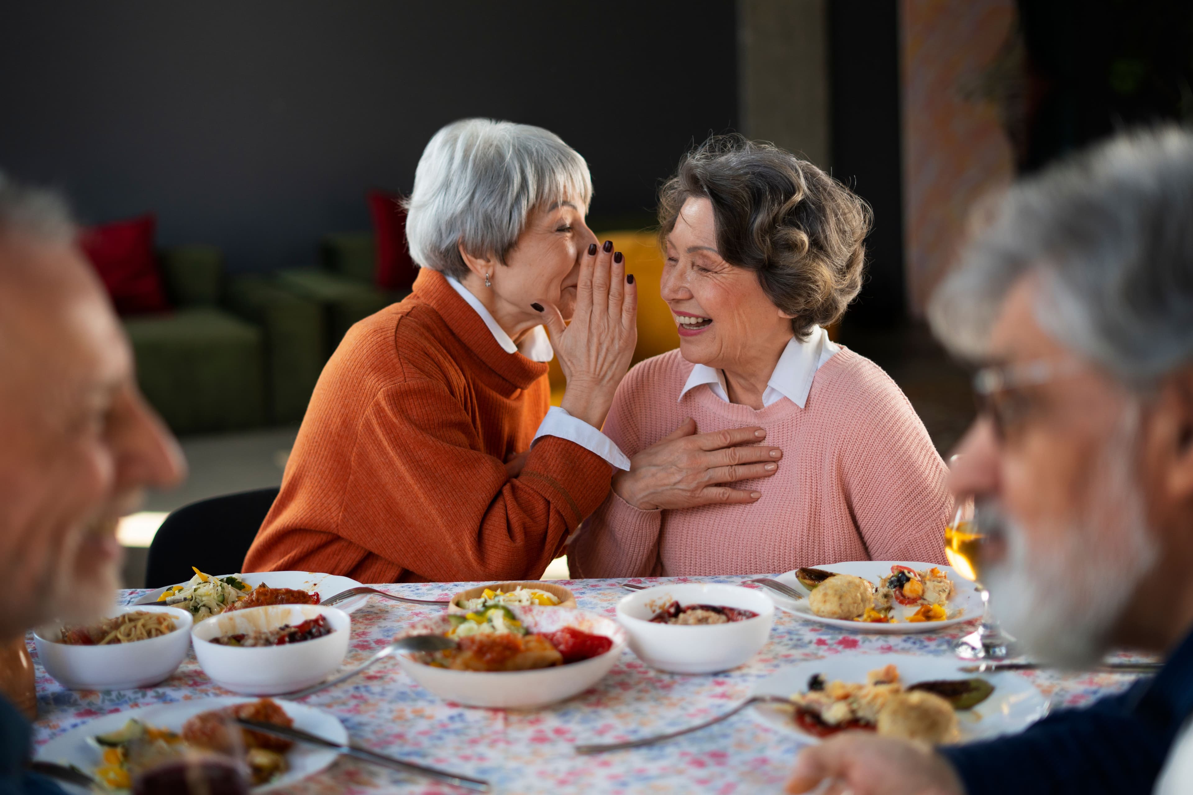 two elderly women are whispering into each other 's ears while sitting at a table with food .