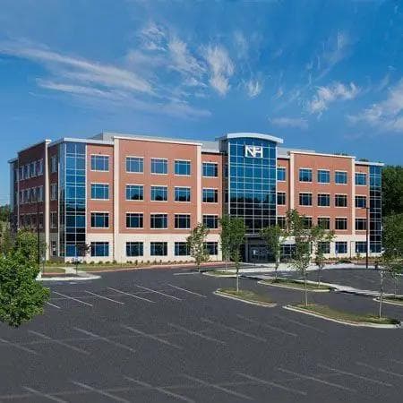 A modern brick and glass building with an NH logo, a large parking lot, and blue sky.