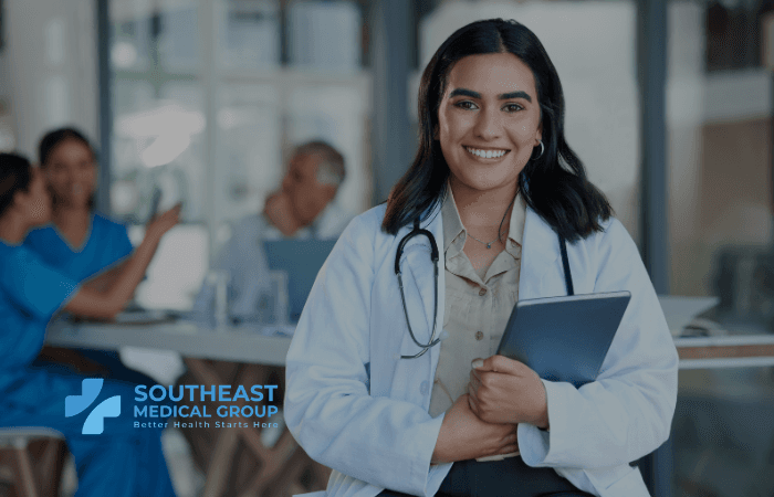 A smiling doctor with a stethoscope holds a tablet, with blurred medical professionals in the background and a blue logo.