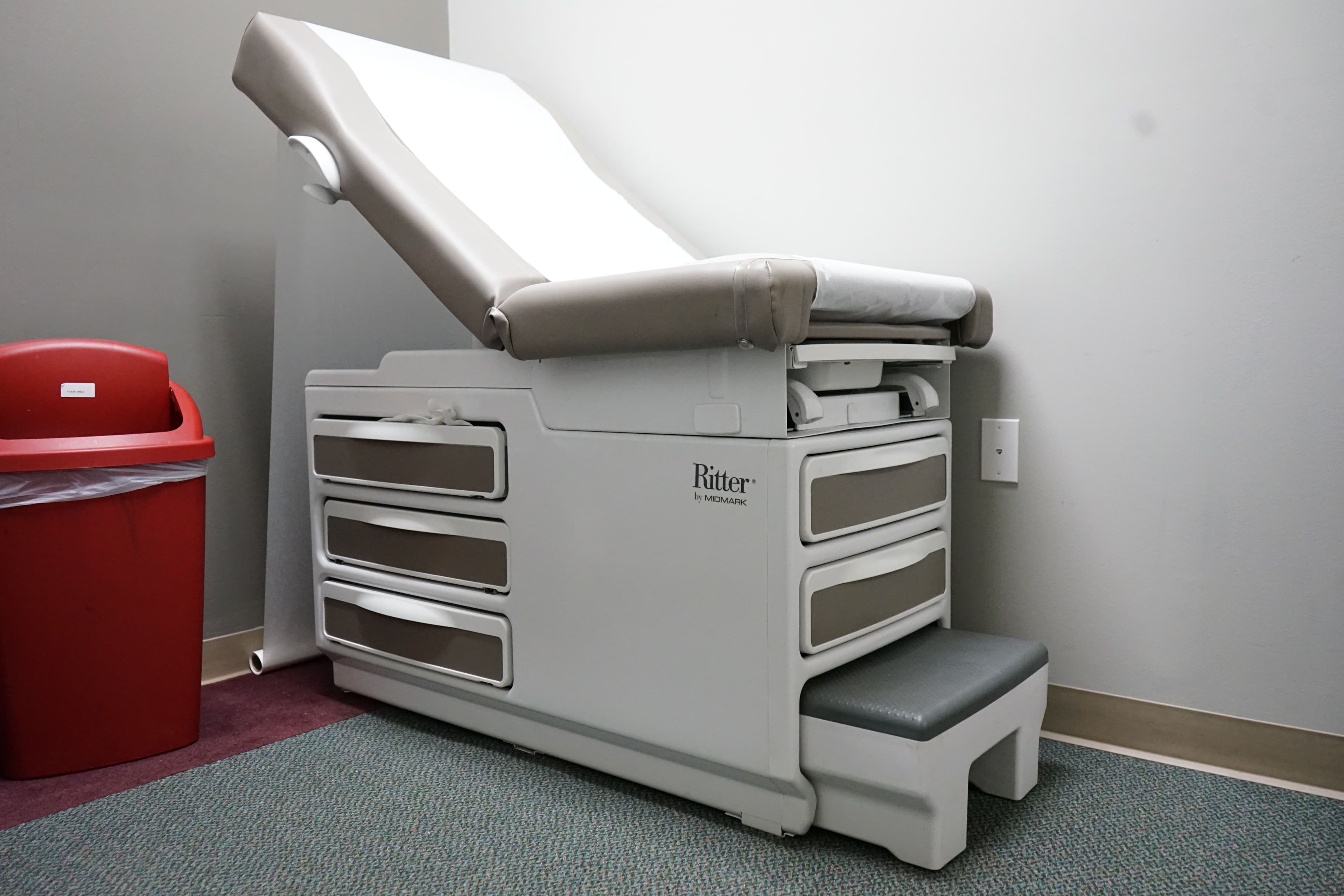 a doctor 's table is sitting in a room next to a red trash can .