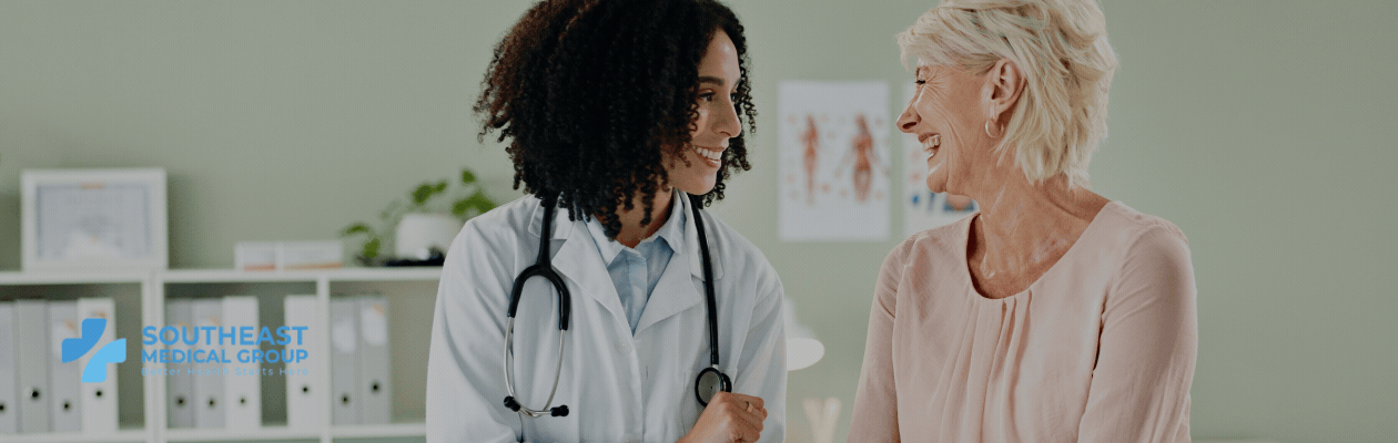 A smiling female doctor talks with an older female patient in a medical office. The Southeast Medical Group logo is visible.