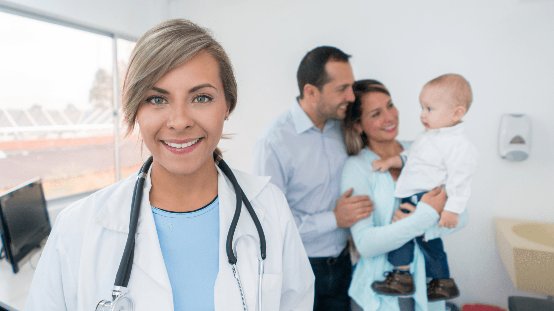 A smiling female doctor with a stethoscope, with a family holding a baby in the background.