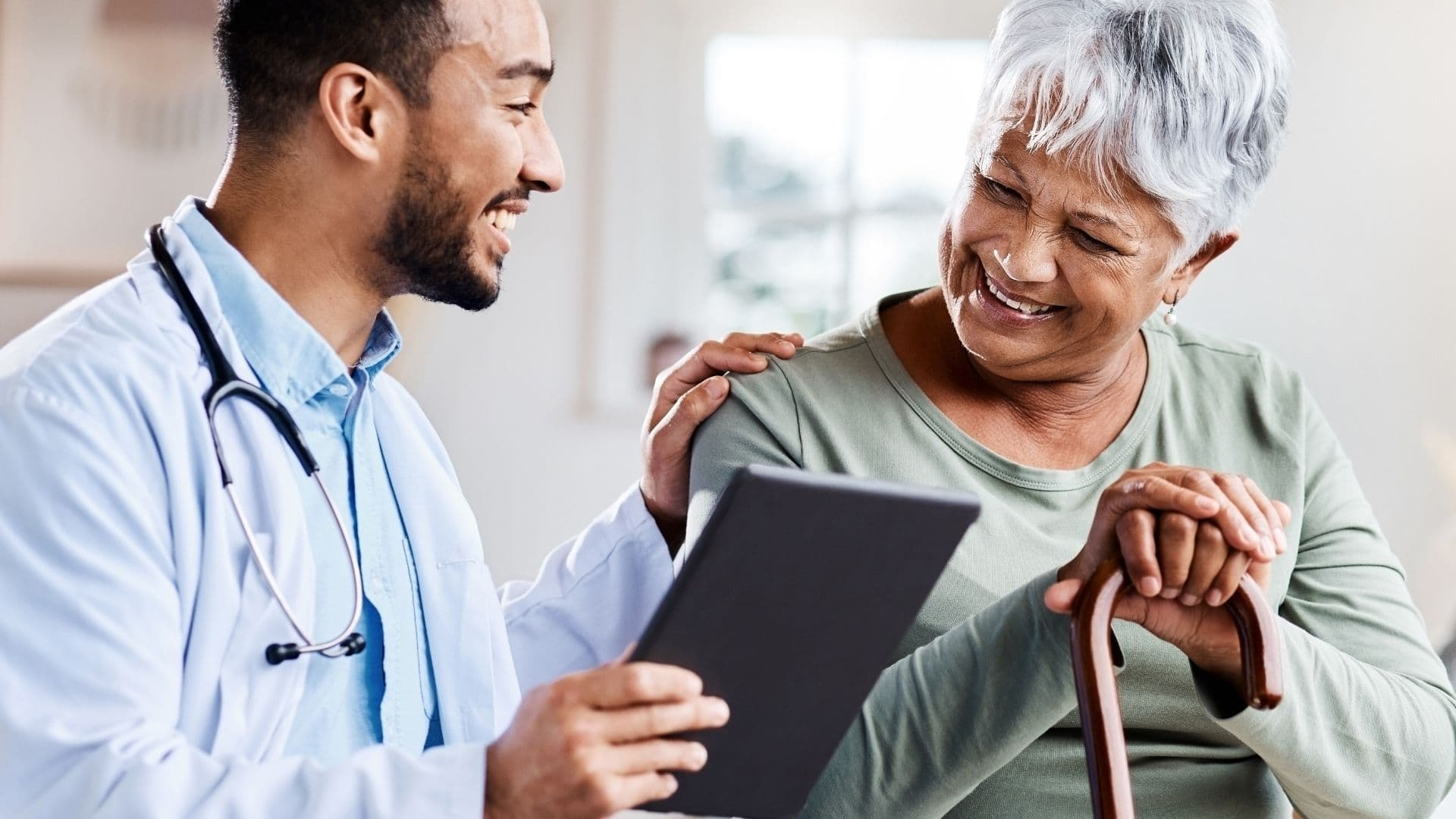 A smiling doctor shows a tablet to an elderly woman with grey hair, who is also smiling and holding a cane.