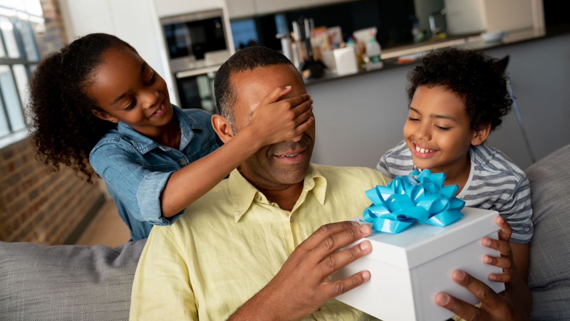 Father's Day Celebration 2024: A man is sitting on a couch receiving a gift from two children .