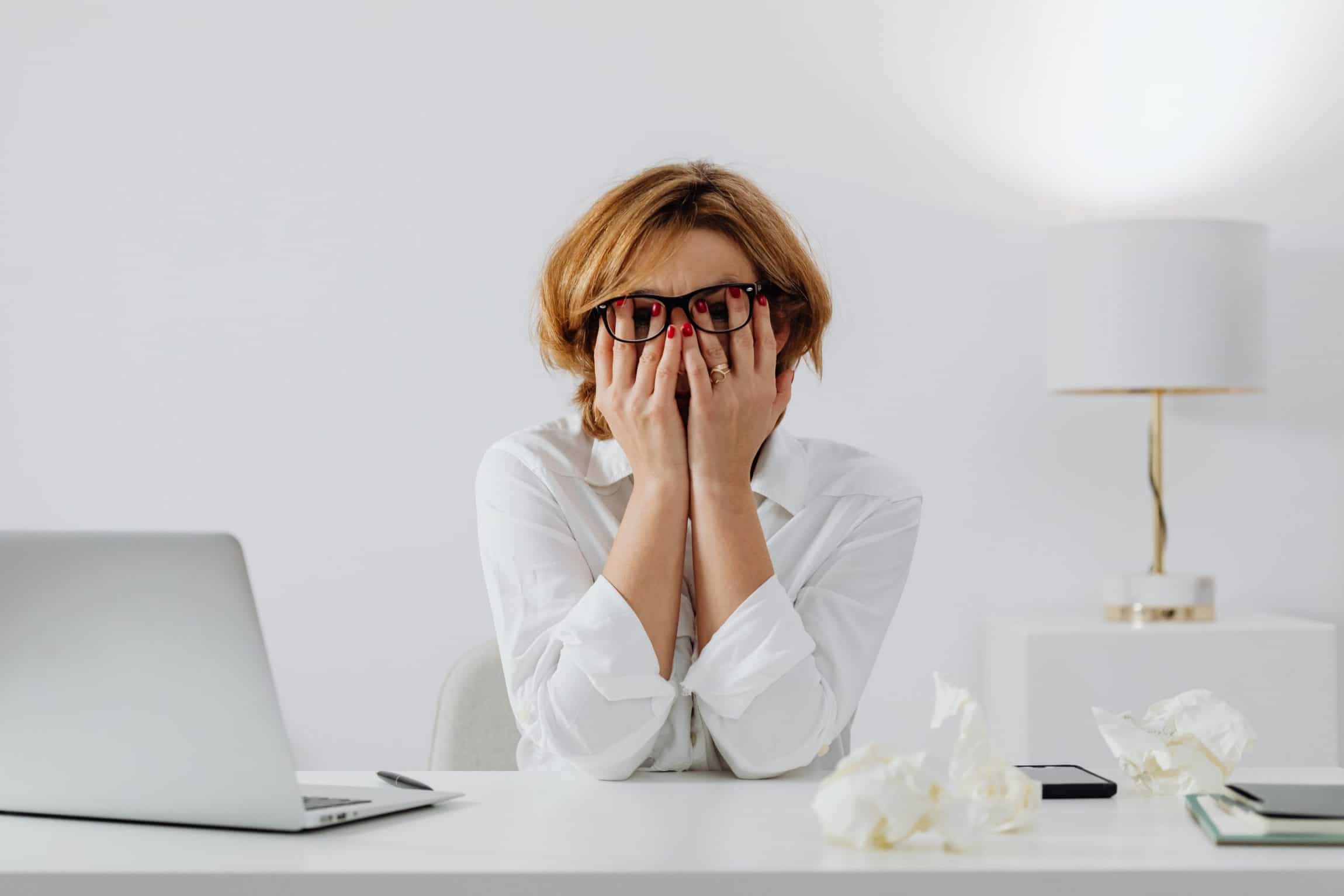 a woman is sitting at a desk with a laptop and covering her face with her hands .