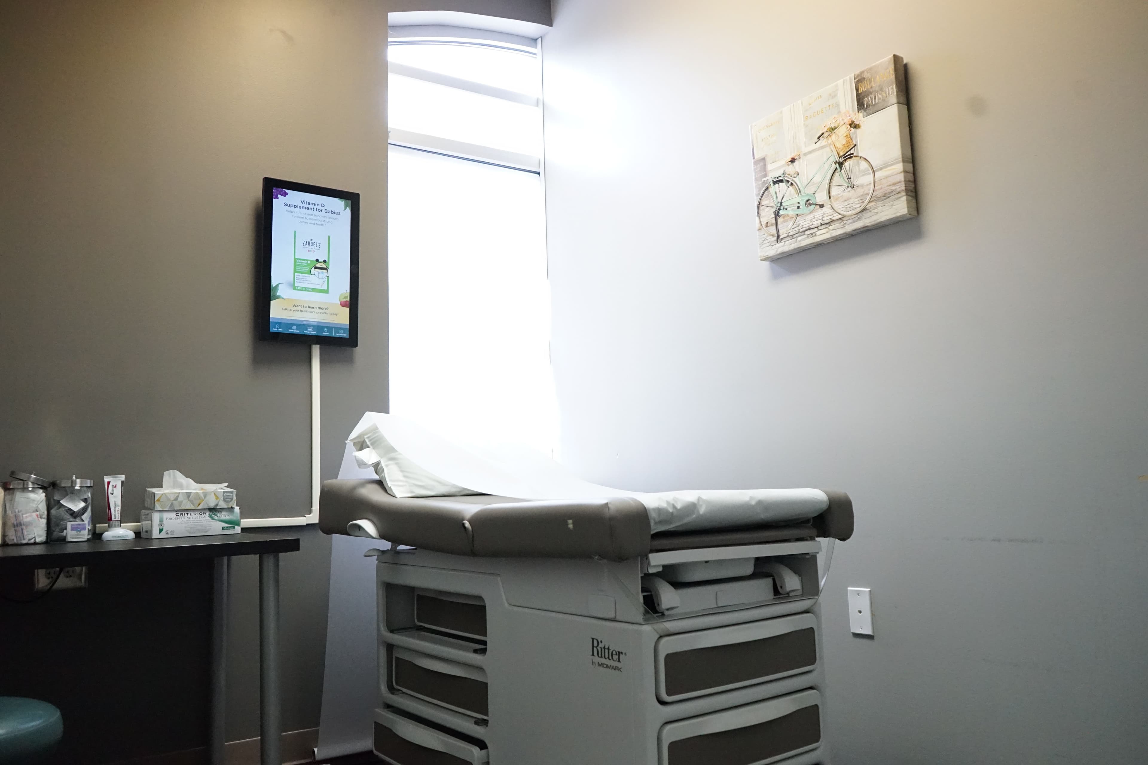 a doctor 's office with a bed , desk , monitor and bicycle painting on the wall .