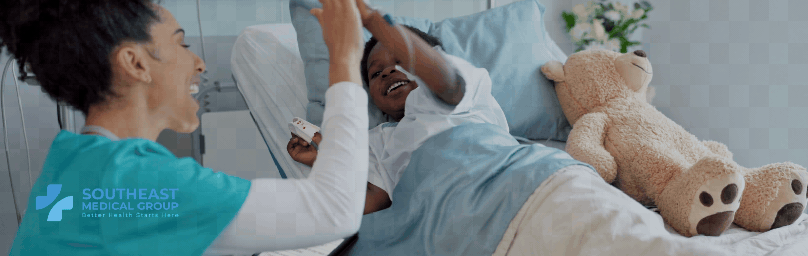 A nurse high-fives a smiling child in a hospital bed with a teddy bear.