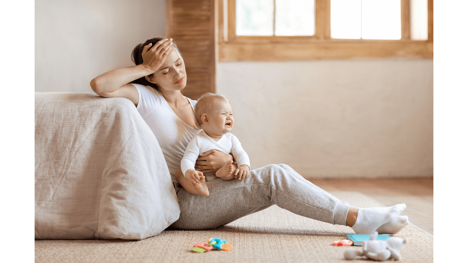 An exhausted mother sits on the floor, hand on her forehead, holding a crying baby.