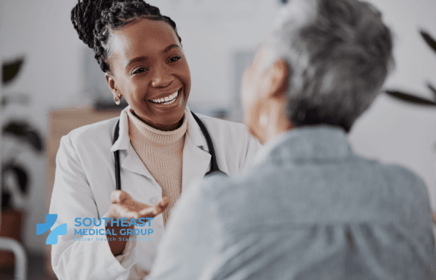 A smiling Black female doctor in a white coat speaks with an older patient whose back is to the camera.