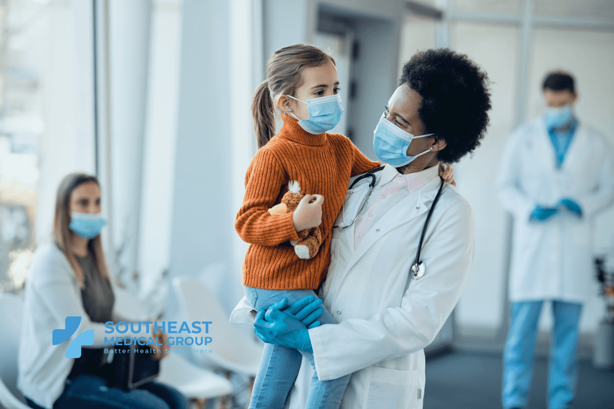 A masked doctor holds a masked young girl with a toy in a medical waiting room.