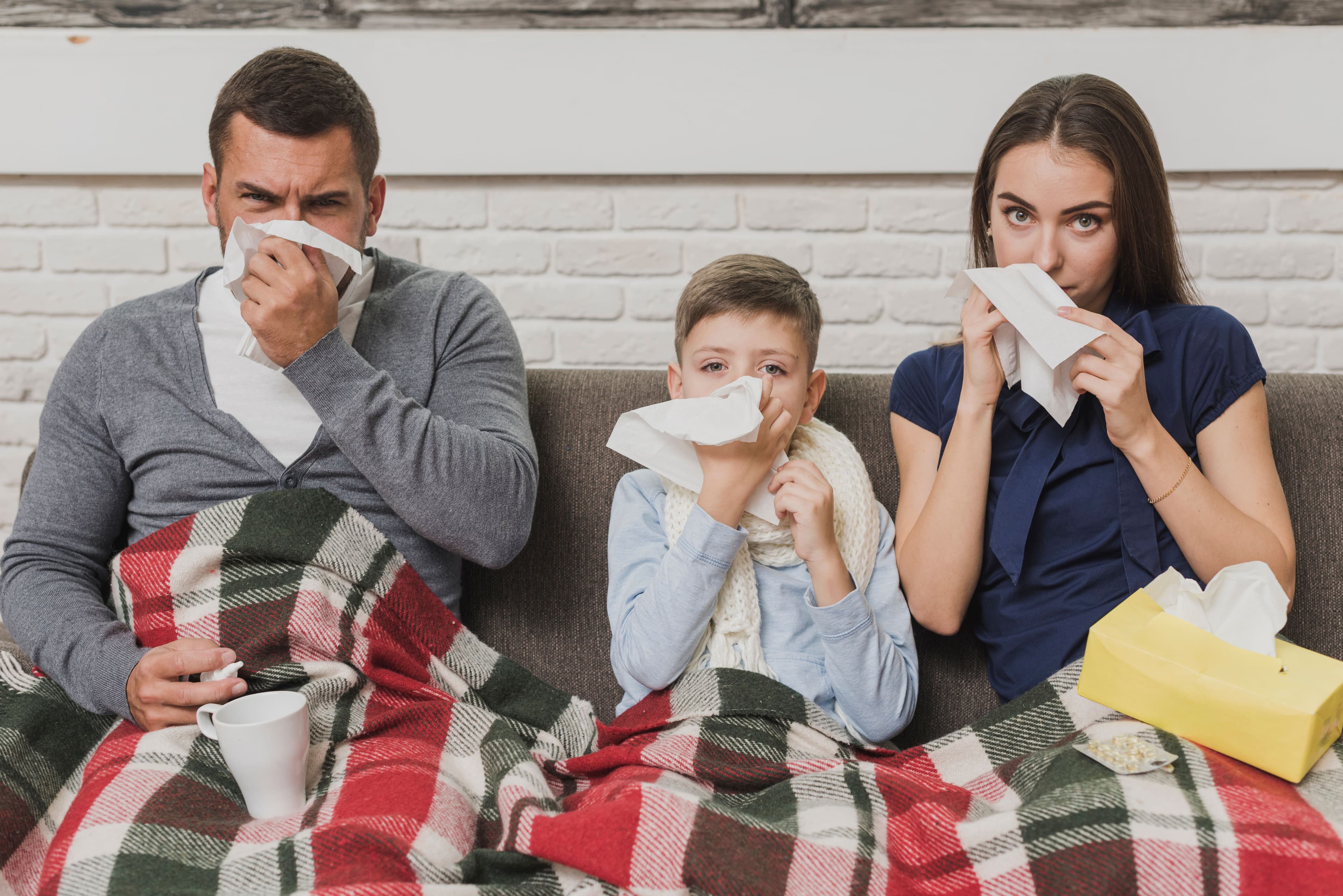 Flu - a family is sitting on a couch blowing their noses with tissues .
